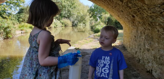 Two young children assess the colour of some canal water, beneath a bridge.