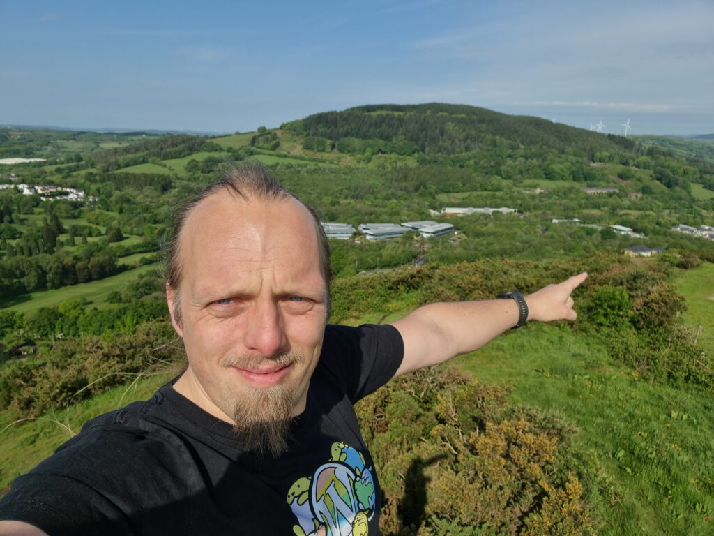 Dan, on a hilltop, points down into the valley between this and the next hill over. Where he's pointing is a white-and-brown-brick building with a grey slate roof in the iconic shape of a Travelodge hotel.