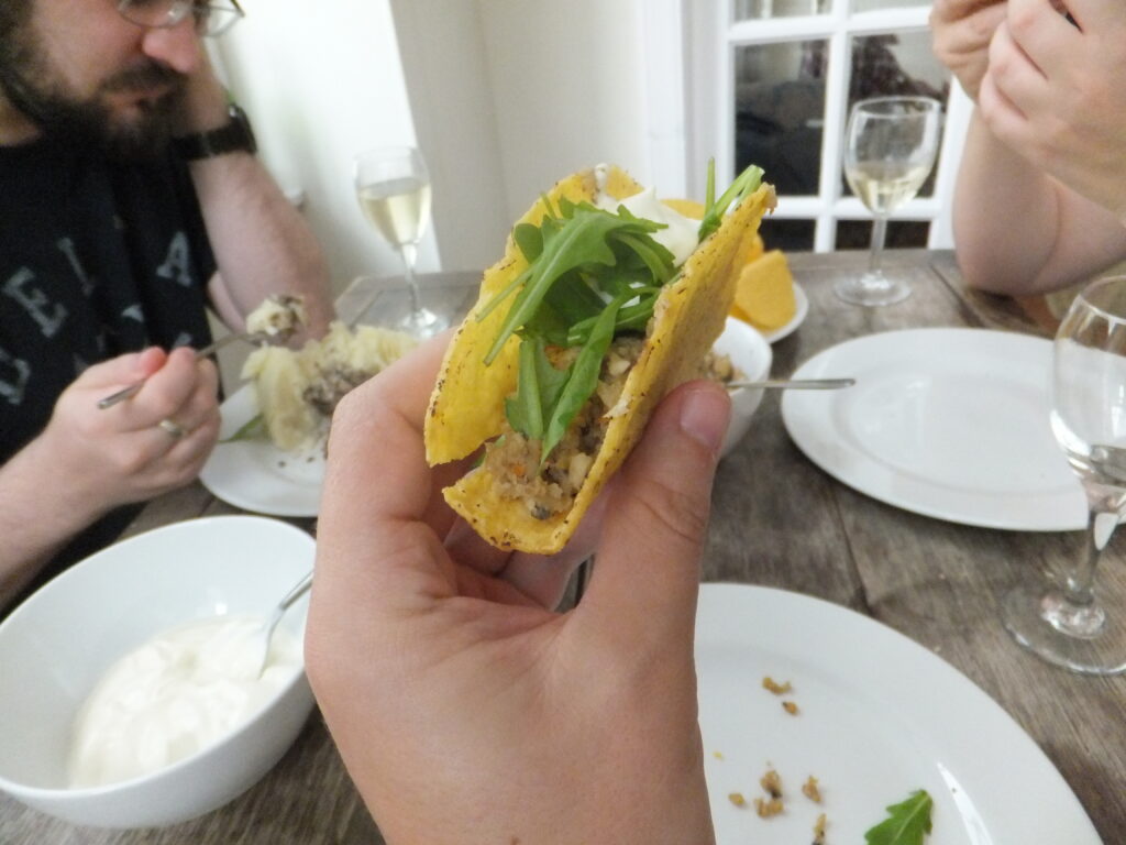 A hand holds a crisp taco containing haggis, mashed potato, rocket, and a blob of sour cream. In the background, JTA can be seen eating his dinner in a more-conventional way: off a plate. There are glasses of wine on the table.