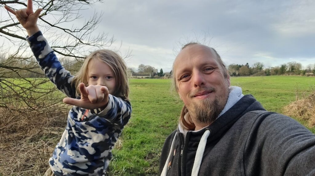 Dan, wearing a grey hoodie, stands in a meadow alongside a boy who's throwing "devil horns" hand signs.