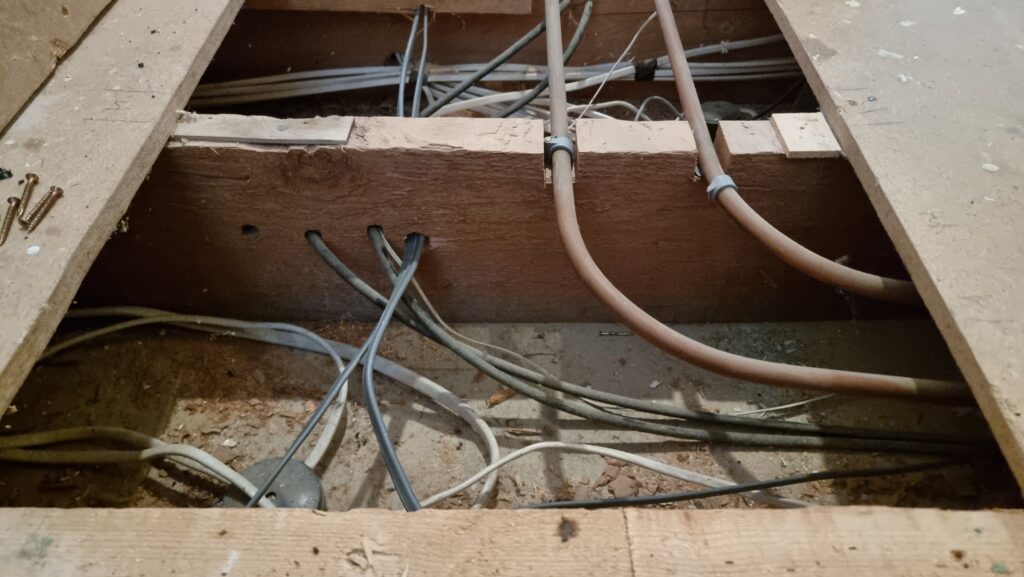 Lifted floor showing central heating pipes and a tangle of electrical cables.