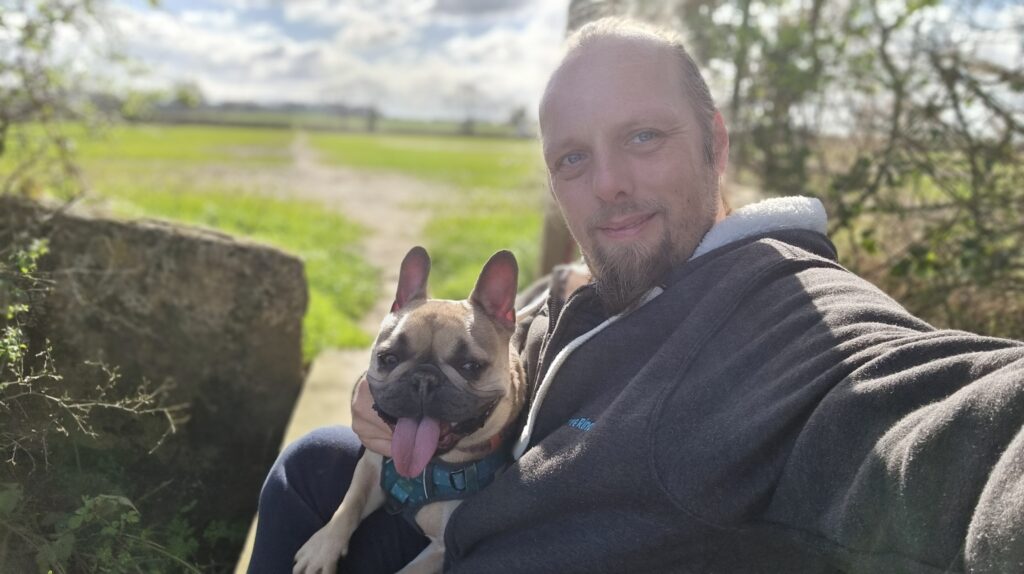 Dan sits on a footbridge in a field, a French bulldog on his lap, in spring morning sunshine.