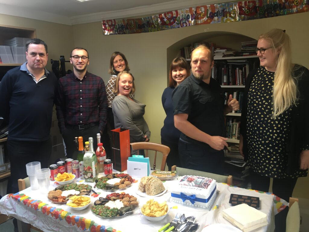 Dan poses with Bodleian coworkers in front of a party feast in a low-ceilinged office.