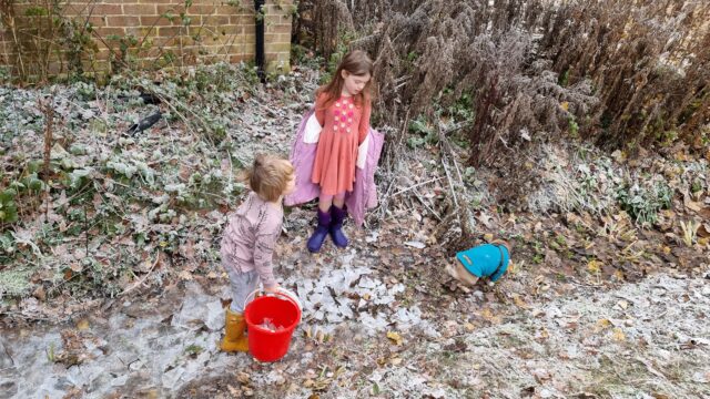 Two children wearing wellies stand in a ditch, breaking ice into chunks.