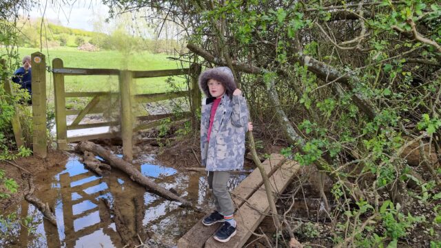 A 7-year-old boy in a grey camo coat balances on a blank over a large muddy puddle: he's about to attempt to cross a log to a gate into the next field (which also looks pretty wet). Ruth, who doesn't much like featuring in photos, has been digitally-removed from this one (she was standing at the far side ready to catch the balancing child!).