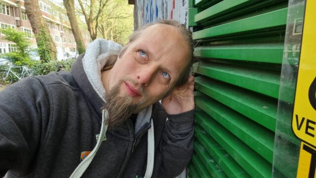 Dan, his hand cupped to his ear, listens closely at the green-painted door of a water pumping station.
