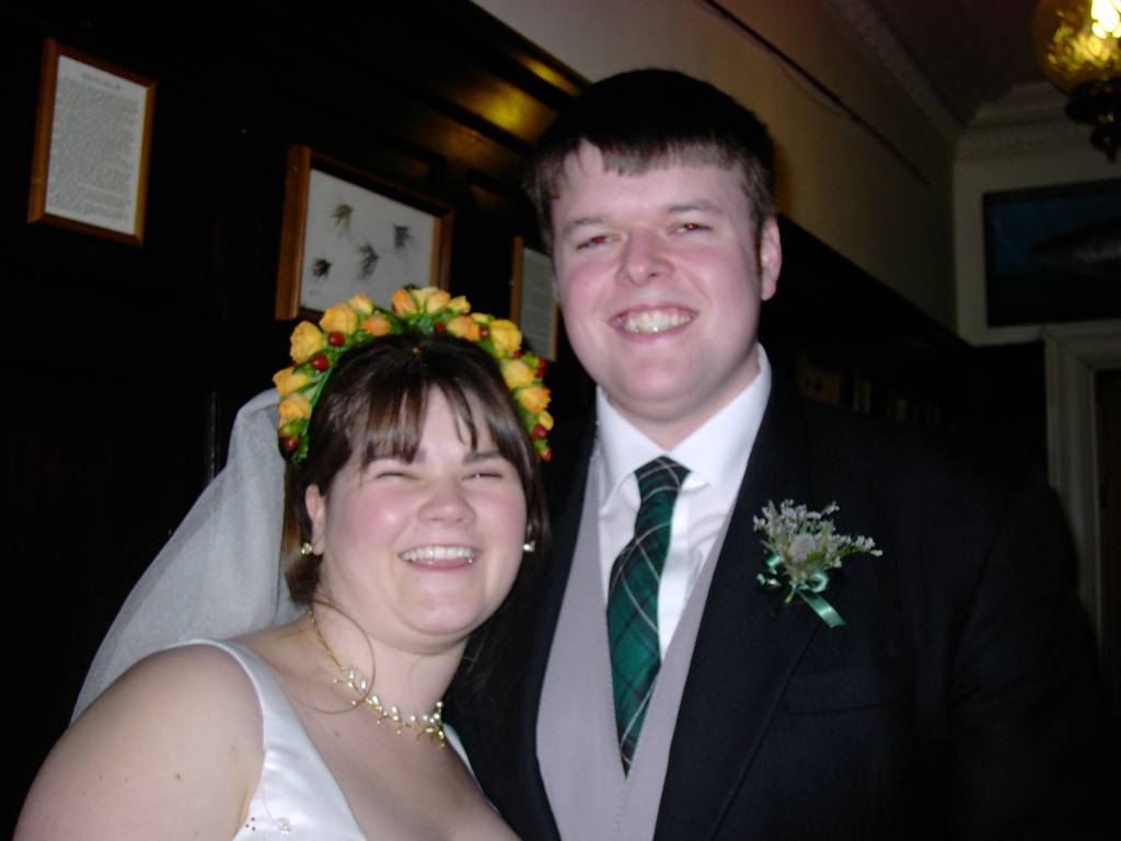 A white couple, bride and groom; she's wearing a white dress and flowers in her hair; he's in a suit with a grey waistcoat and a thistle buttonhole.