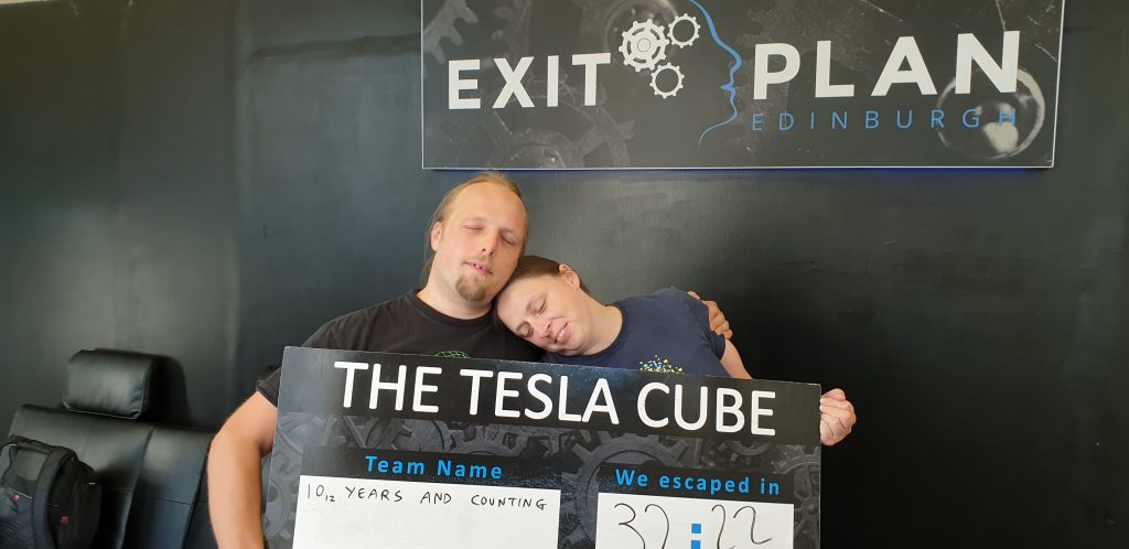 Dan and Ruth pretend to be asleep while holding a sign that says that they solved The Tesla Cube escape room in 32 mintues and 22 seconds.