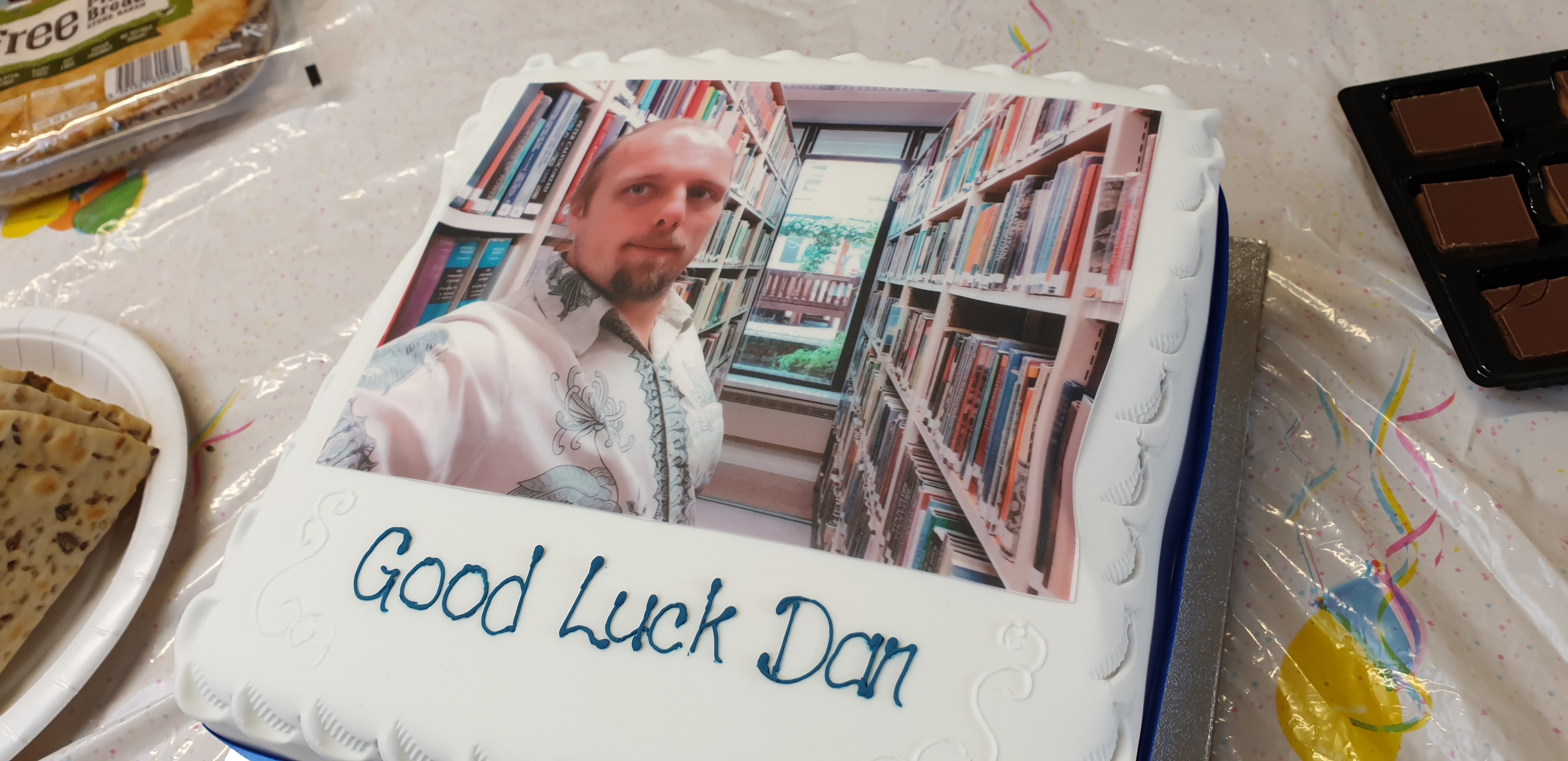 A cake with icing printed with a picture of Dan in a library. Beneath are iced the words "Good Luck Dan".