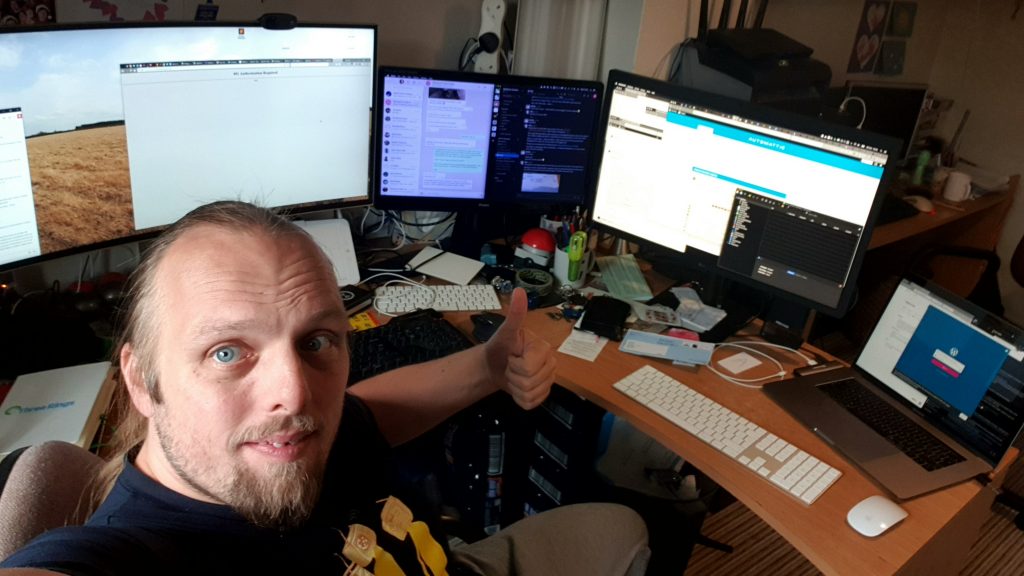 Dan in front of a crowded desk: two large monitors, a regular-sized monitor, and a laptop are visible, among clutter.