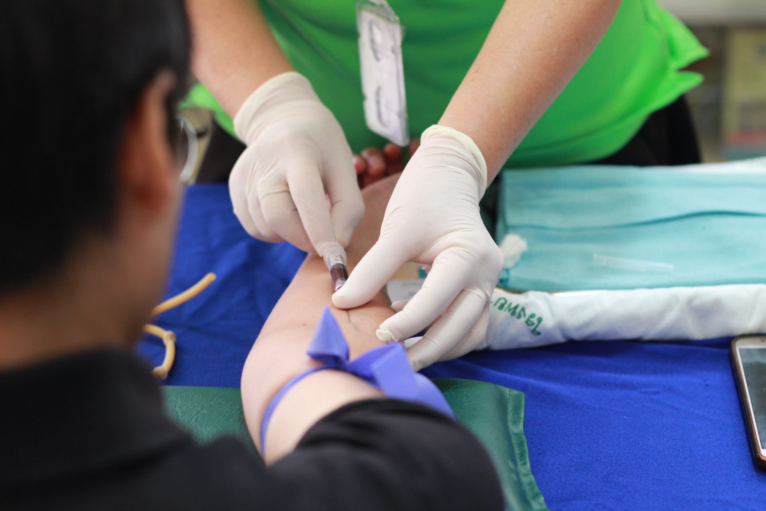 A nurse wearing gloves uses a hyperdermic needle to take a blood sample from a patients' arm, as seen from over the patient's shoulder.