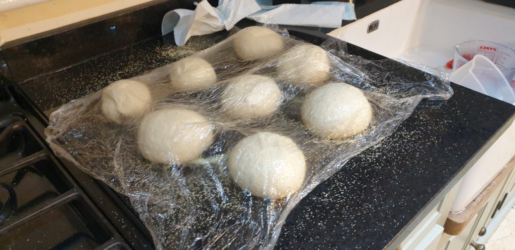 Pizza dough balls under cling film on a black marble surface.