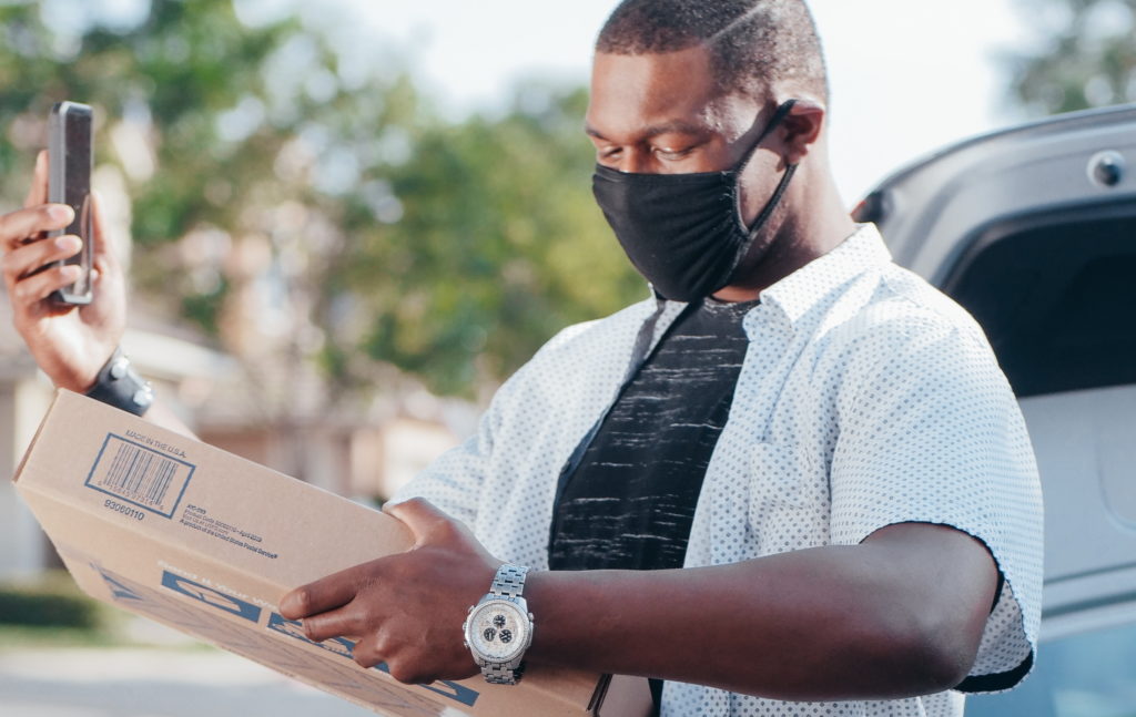 Delivery man, wearing a face mask, holding a parcel and checking his mobile phone. Photo by Kindel Media from Pexels.