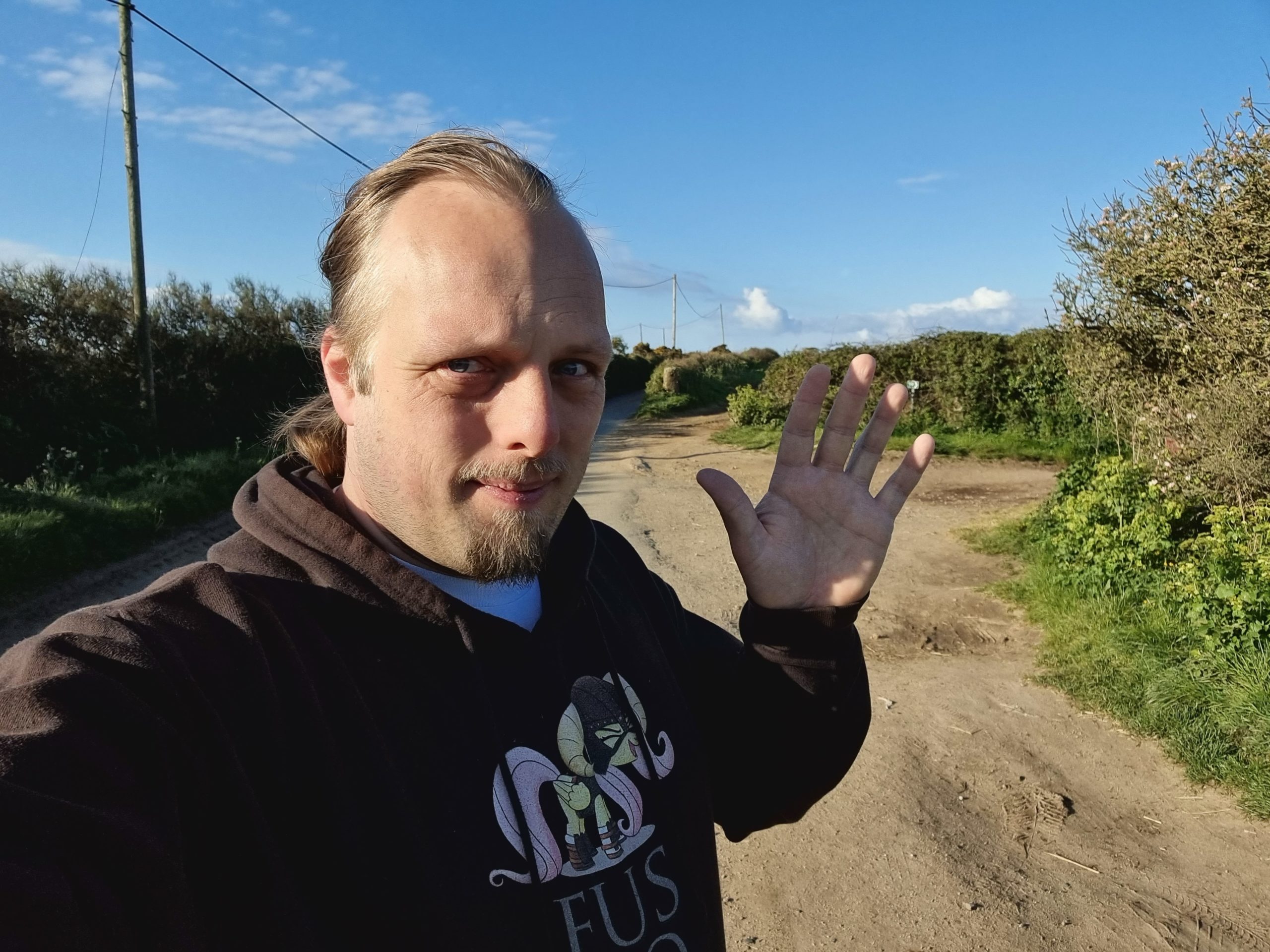 Dan, on a country lane in Cornwall, in front of a bright blue sky, waves to the camera.