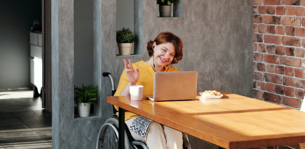 A woman in a wheelchair waves to a colleague via her laptop screen; she's smiling and has a cup of coffee by her side. Photo by Marcus Aurelius from Pexels.