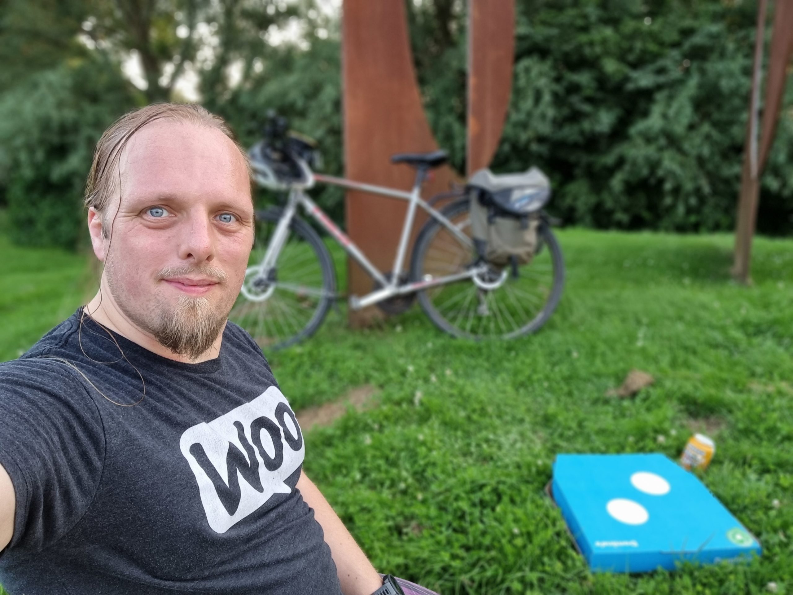 Dan sits on the grass with a Dominos pizza box and a can of beer. His bike is in the background, leaning on a stylistically-corroded piece of metal artwork.