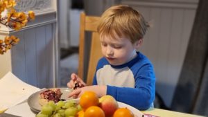 Boy eating a crumpet alongside a bowl of fruit.