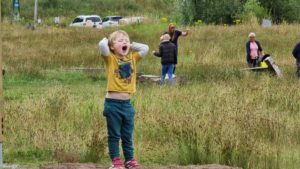 A boy shouts from the top of a grassy knoll near a car park.