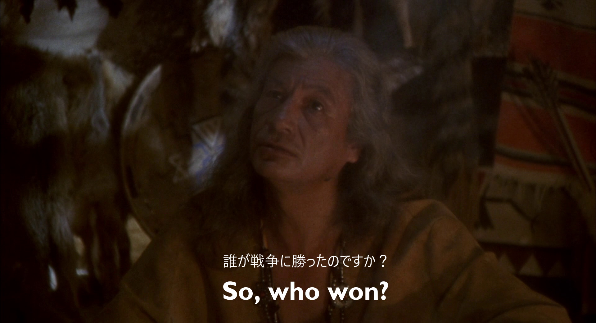 Still from Hot Shots!. Owatonna, an older Native American man, sits surrounded by animal skins. An English subtitle reads "So, who won?" A Japanese subtitle reads "誰が戦争に勝ったのですか?", which translates as "Who won the war?"