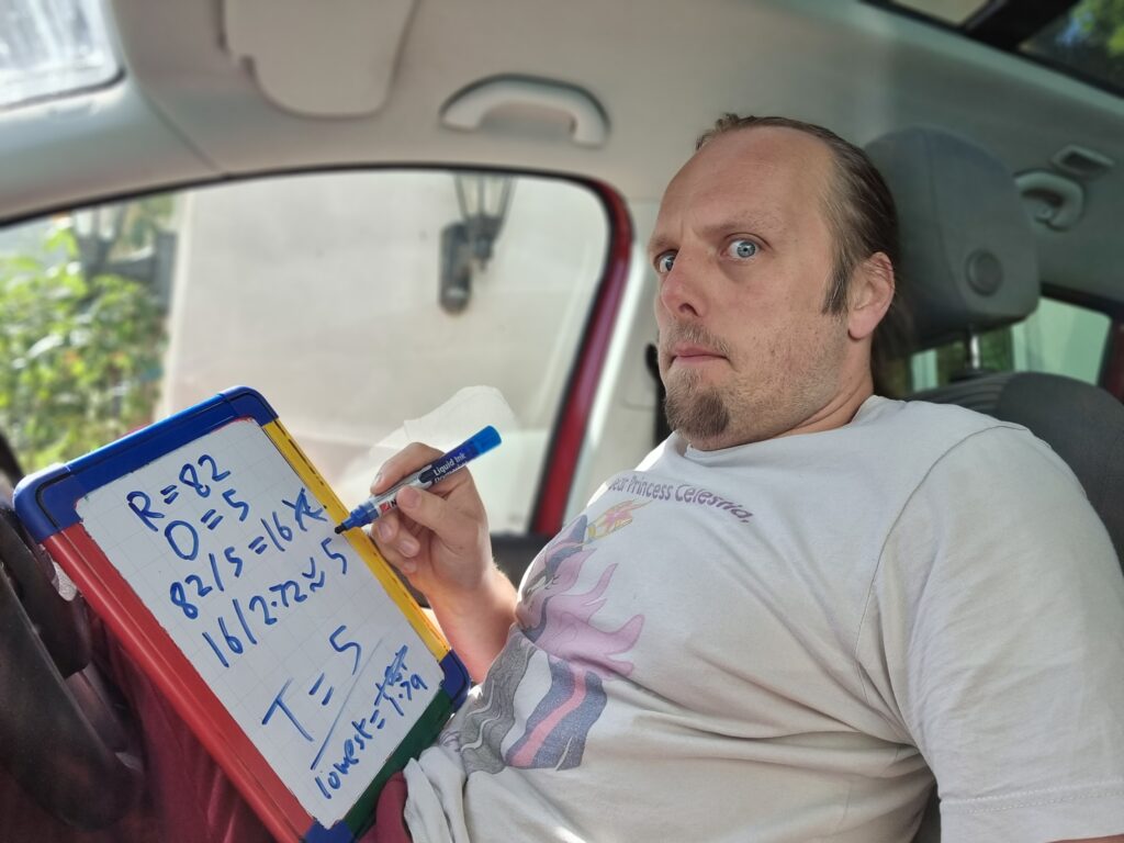 Dan sitting in the driving seat of a car, doing maths on a portable whiteboard.