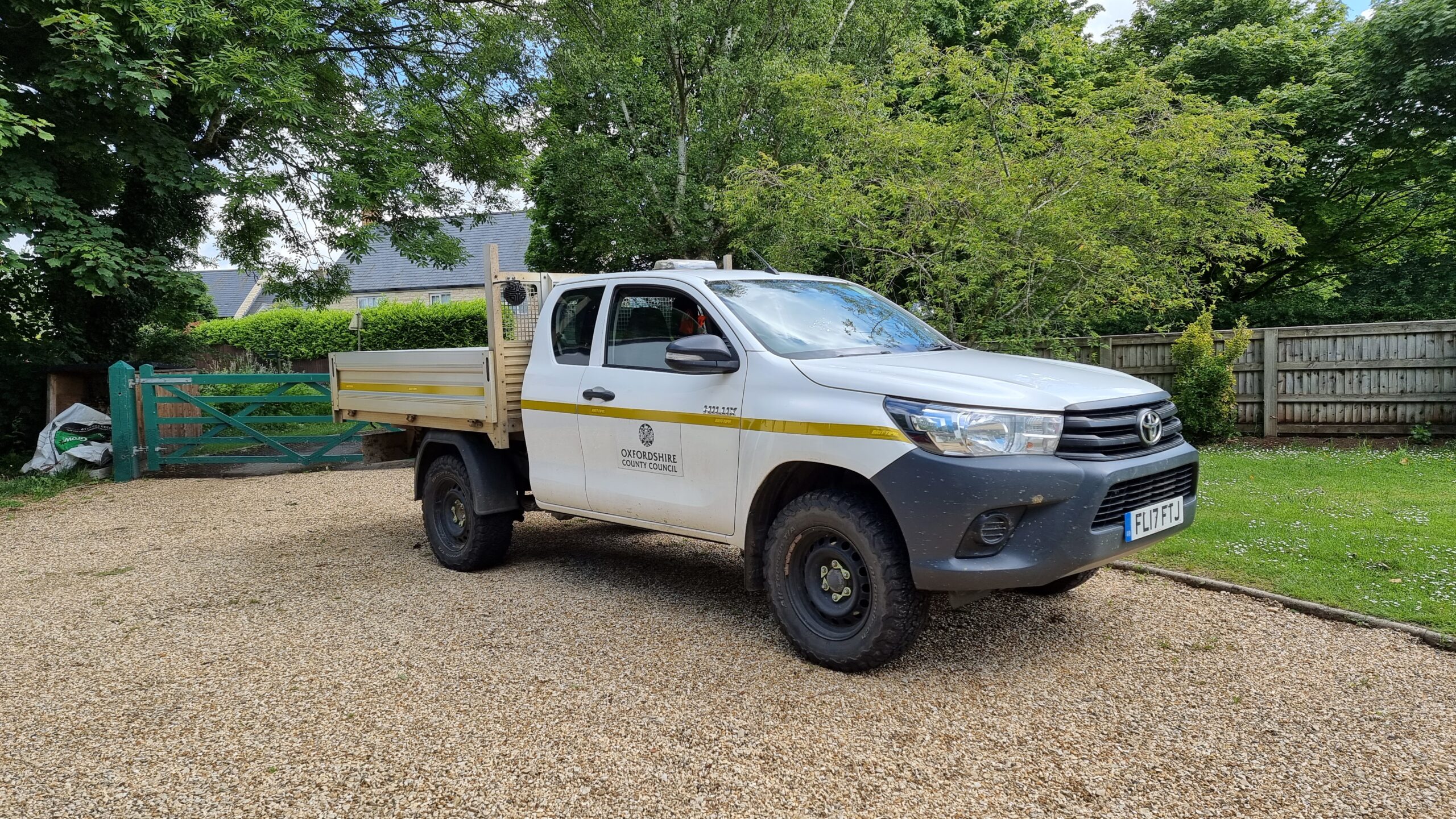 A rugged white flatbed van with a yellow stripe and the logo of Oxfordshire County Council sits on a gravel driveway in a garden. The sun glistens off the windscreen and the driver-side window is partially ajar.
