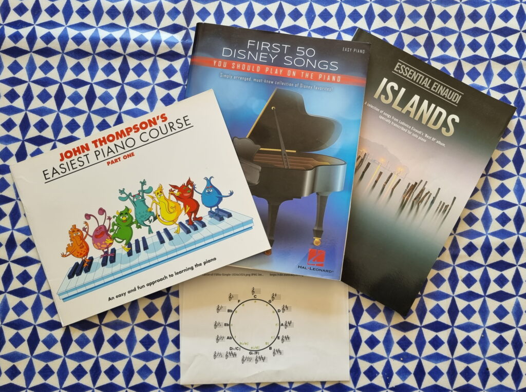 Three books on a blue-and-white tablecloth: John Thompson's Easiest Piano Course (Part One), First 50 Disney Songs, and Essential Einaudi - Islands. Beneath them sits a simplified diagram showing the circle of fifths.