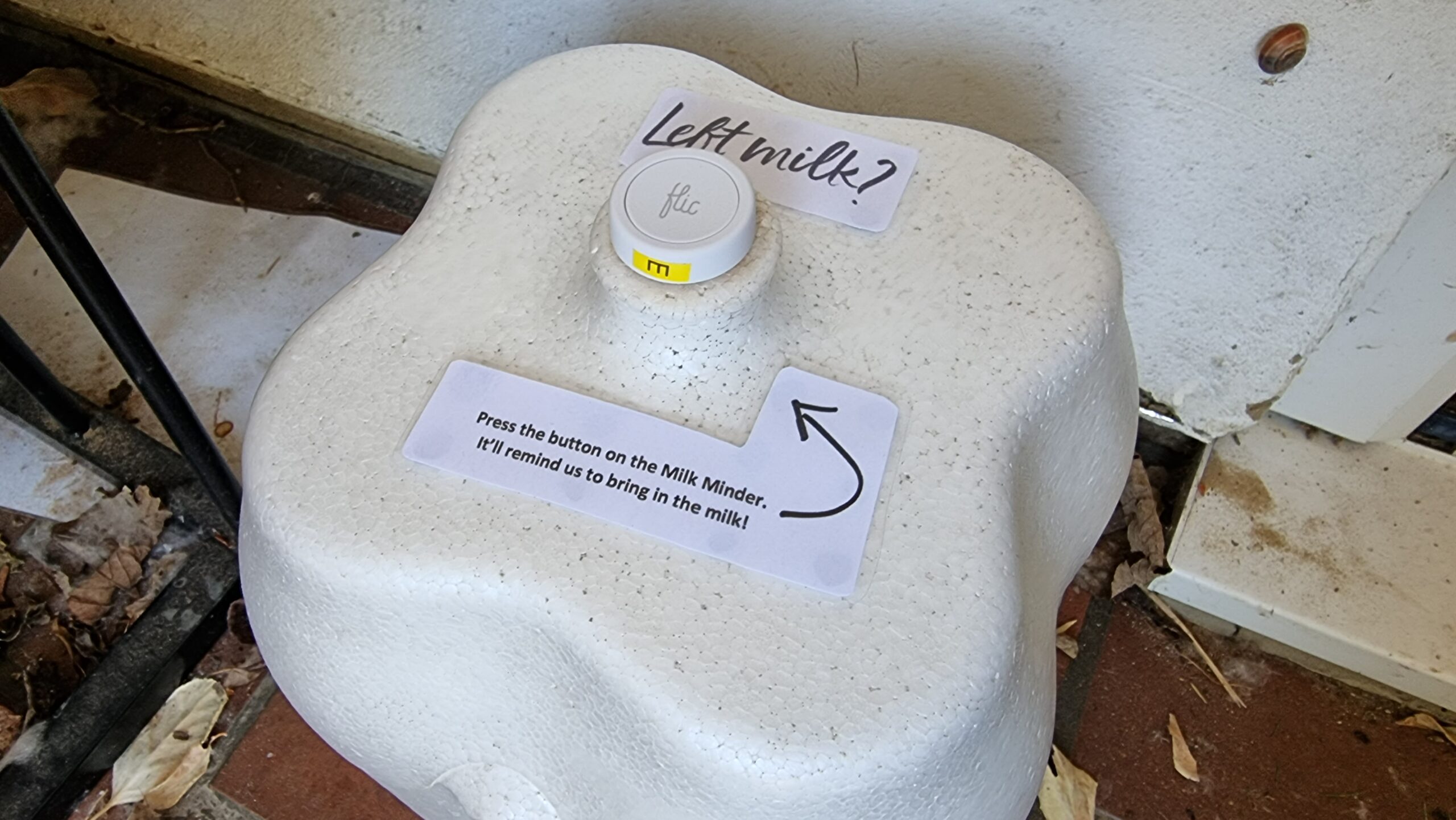 Milk container, with a Flic 2 button attached to the handle of the lid and a laminated notice attached, reading: "Left milk? Press the button on the Milk Minder. It'll remind us to bring in the milk!"