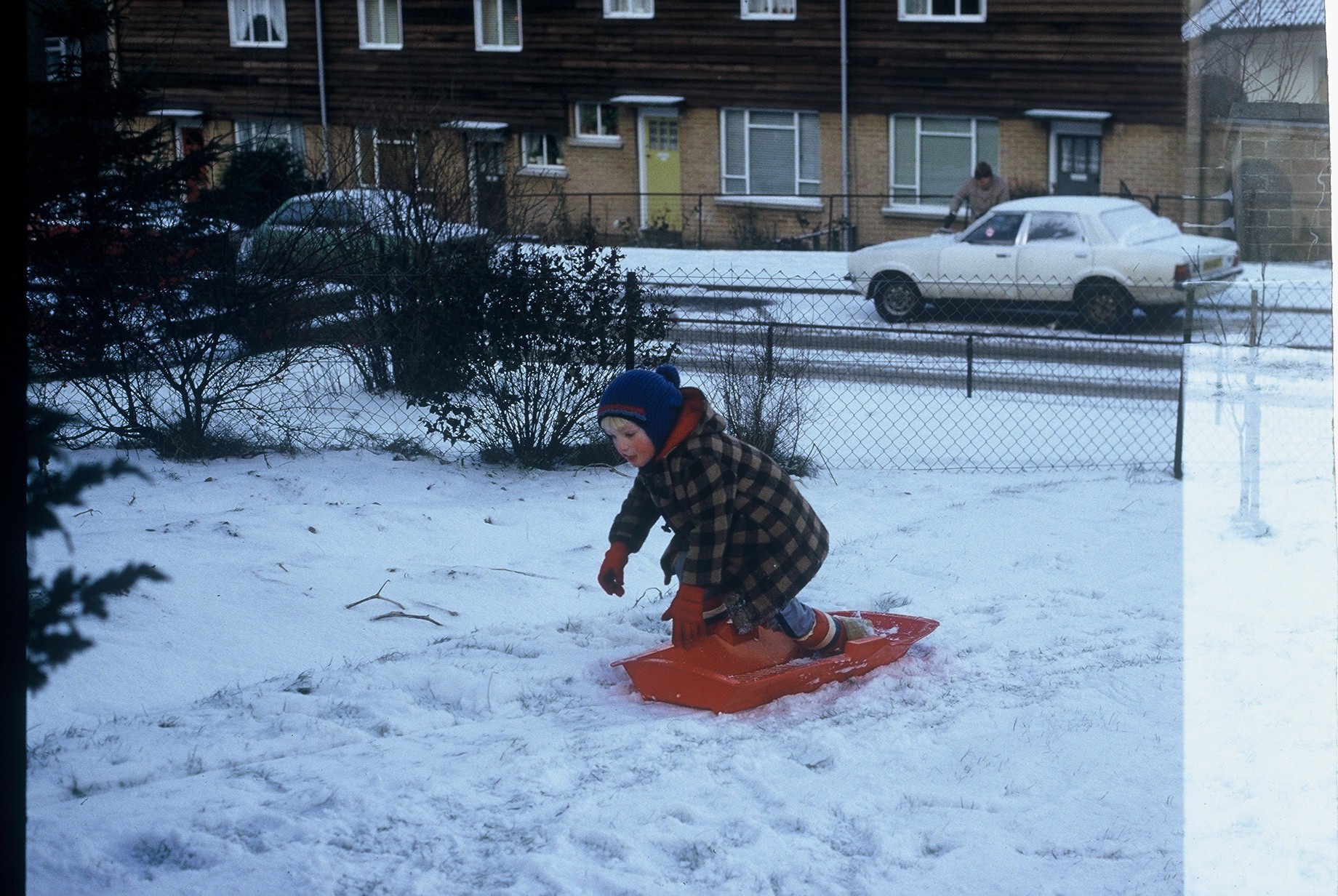 Dan, aged around 4, dressed in a duffel coat, bobble hat and gloves, kneeling on a red plastic sledge in a snow-filled garden. The garden is bordered by a wire fence, and in the background a man can be seen scraping icee off a car.