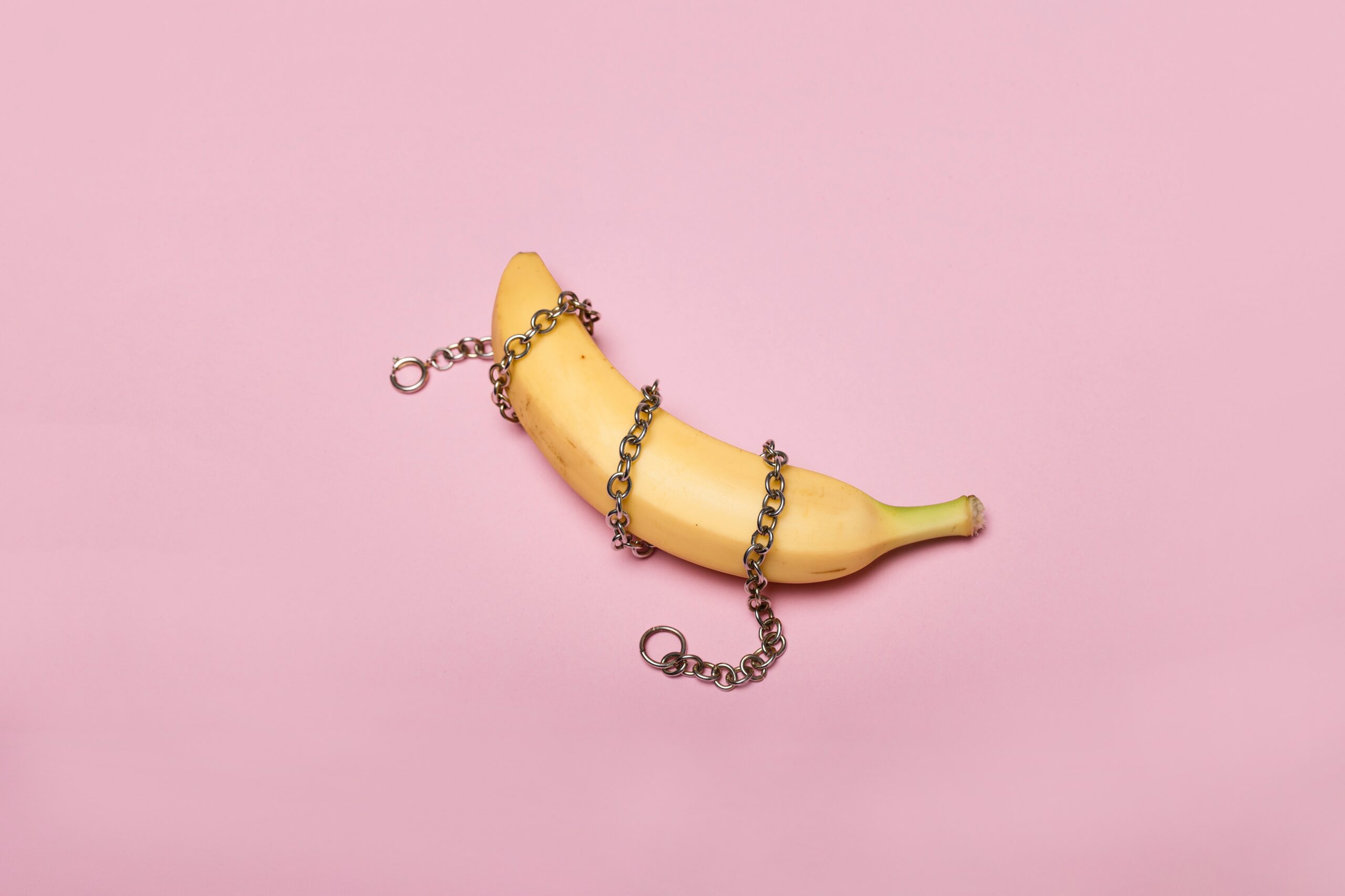 Photograph showing a yellow banana on a pink background. The banana has a silver chain wrapped around it three times. Photo courtesy Deon Black.