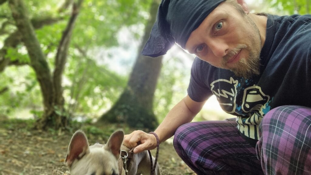 Dan, wearing purple pyjama bottoms, a black t-shirt, and a black bandana, sits in a woodland setting with a dog who's just put herself off the bottom of the frame at the last second.