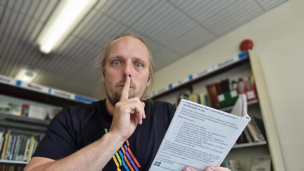 Dan sits in a library, holding a small book and with his finger to his lips in a 'shush' gesture. The back cover of the book contains text identifying it as a geocache.