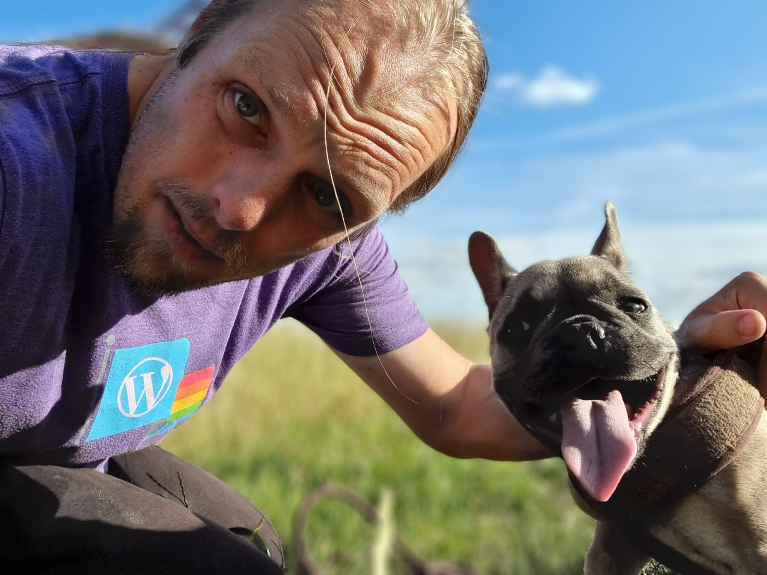 Under bright blue skies and in a green field, Dan and Demmy stare into the camera. Dan's flushed from the heat and wearing a purple-and-rainbow "WordPress Pride" t-shirt; Demmy's tongue hangs low and she's clearly panting.