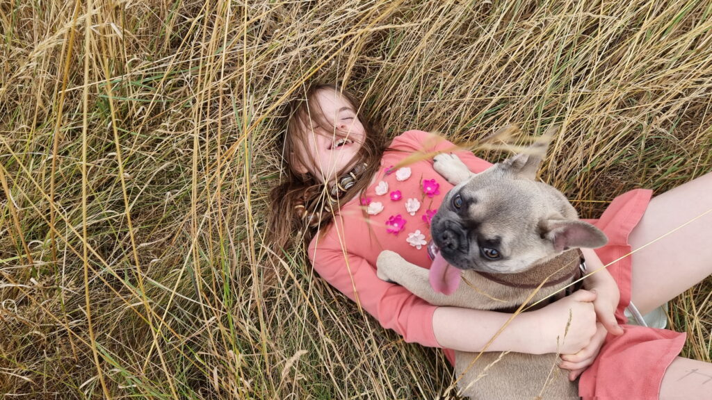 An 8-year-old girl in a flowery pink dress lies on her back in a meadow of long dry grass, laughing with her eyes closed. On her belly, Demmy lies, looking up towards the camera with her tongue hanging out.