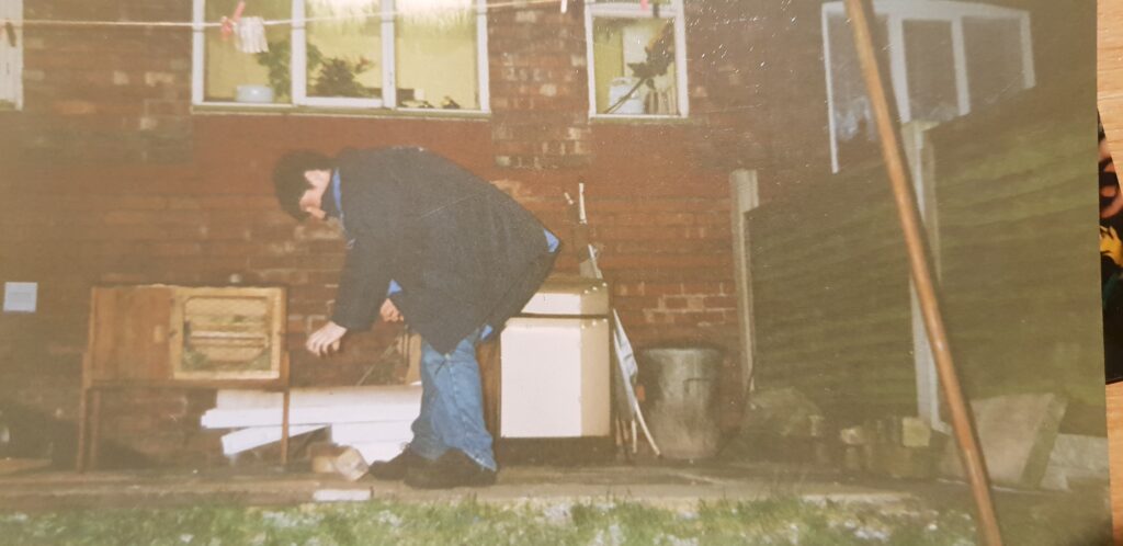 A young man in jeans and a blue coat stands on the patio in the back garden of a terraced house, dropping a half-brick onto the floor. In the background, an unused rabbit hutch and a dustbin can be seen. The photo is clearly taken using a flash, at night.