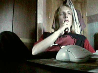 Dan, as a teenager, sits at a desk with his hand to his chin. In the foreground, a beiege two-button wired ball-type computer mouse rests on the corner of the desk. Dan is wearing a black t-shirt with a red devil face printed onto it.