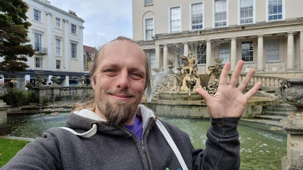 In an urban environment, Dan waves. Behind him, a stone fountain-statue representing Neptune, surrounded by sea creatures, can be seen.