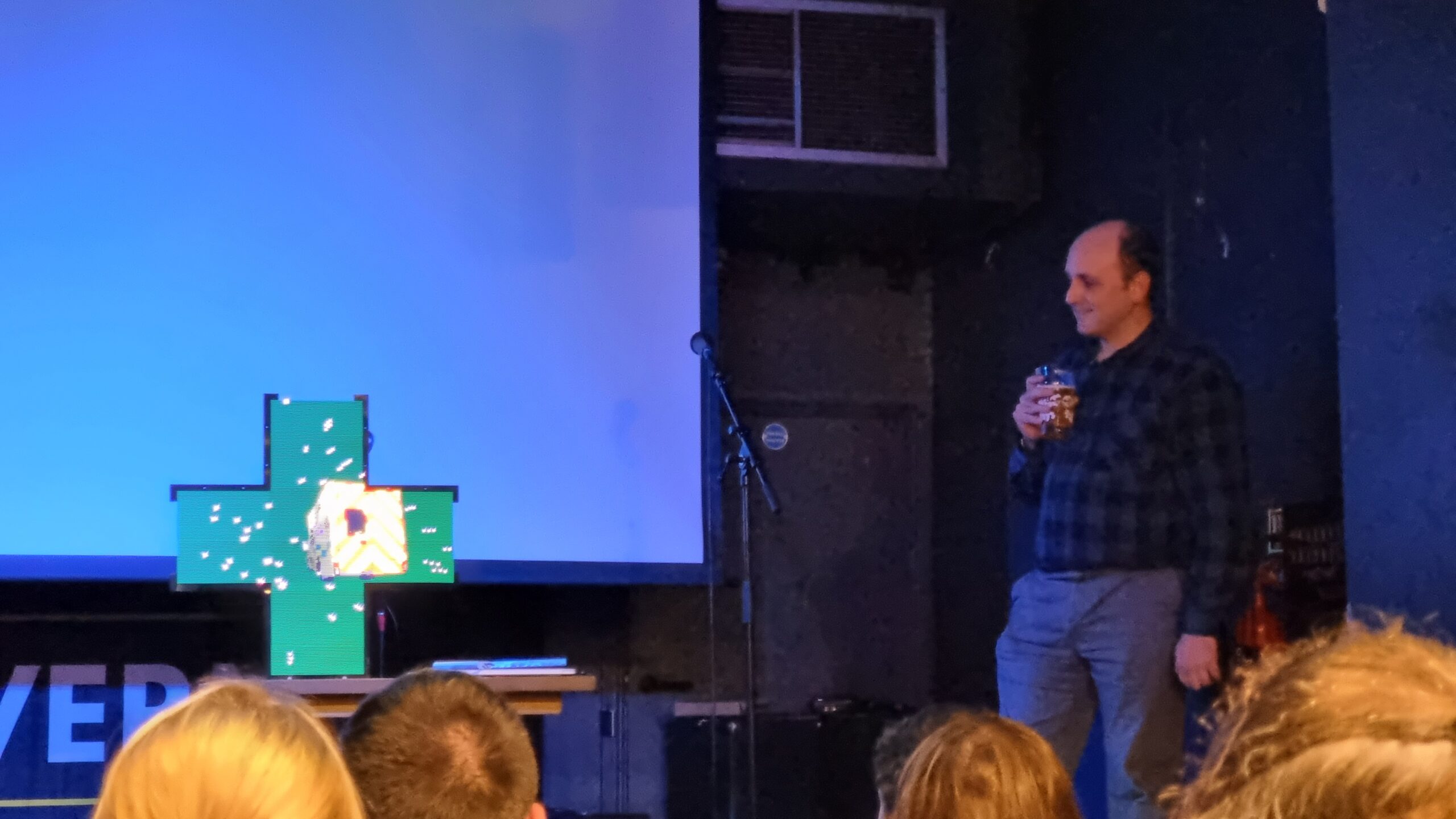 Matt Westcott stands to the side of a stage, drinking beer, while centrestage a cross-shaped "pharmacy sign" projects an animation of an ambulance rocketing into a starfield.