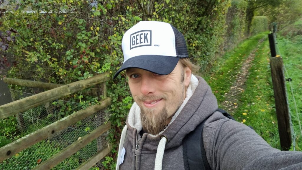 Dan, outdoors on a grassy path, wearing a grey hoodie. On his head is a "trucker cap" emblazoned with the word "GEEK" and, in smaller writing "#OGN52".