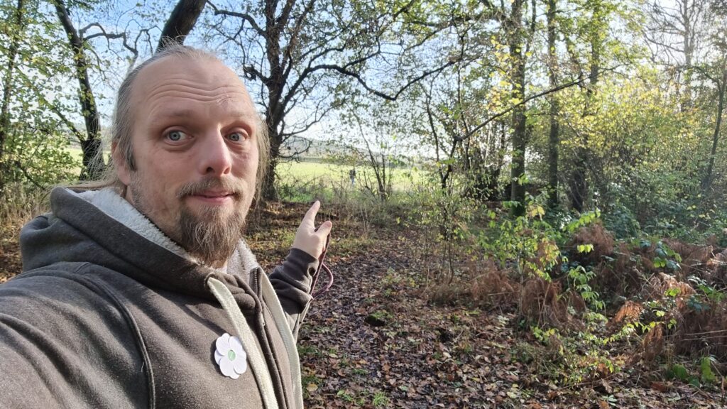 Dan, wearing a grey Three Rings hoodie with a white poppy pinned to it, holding the end of a dog lead, points out of the edge of the woodland he's in towards a humanoid figure in the distance.