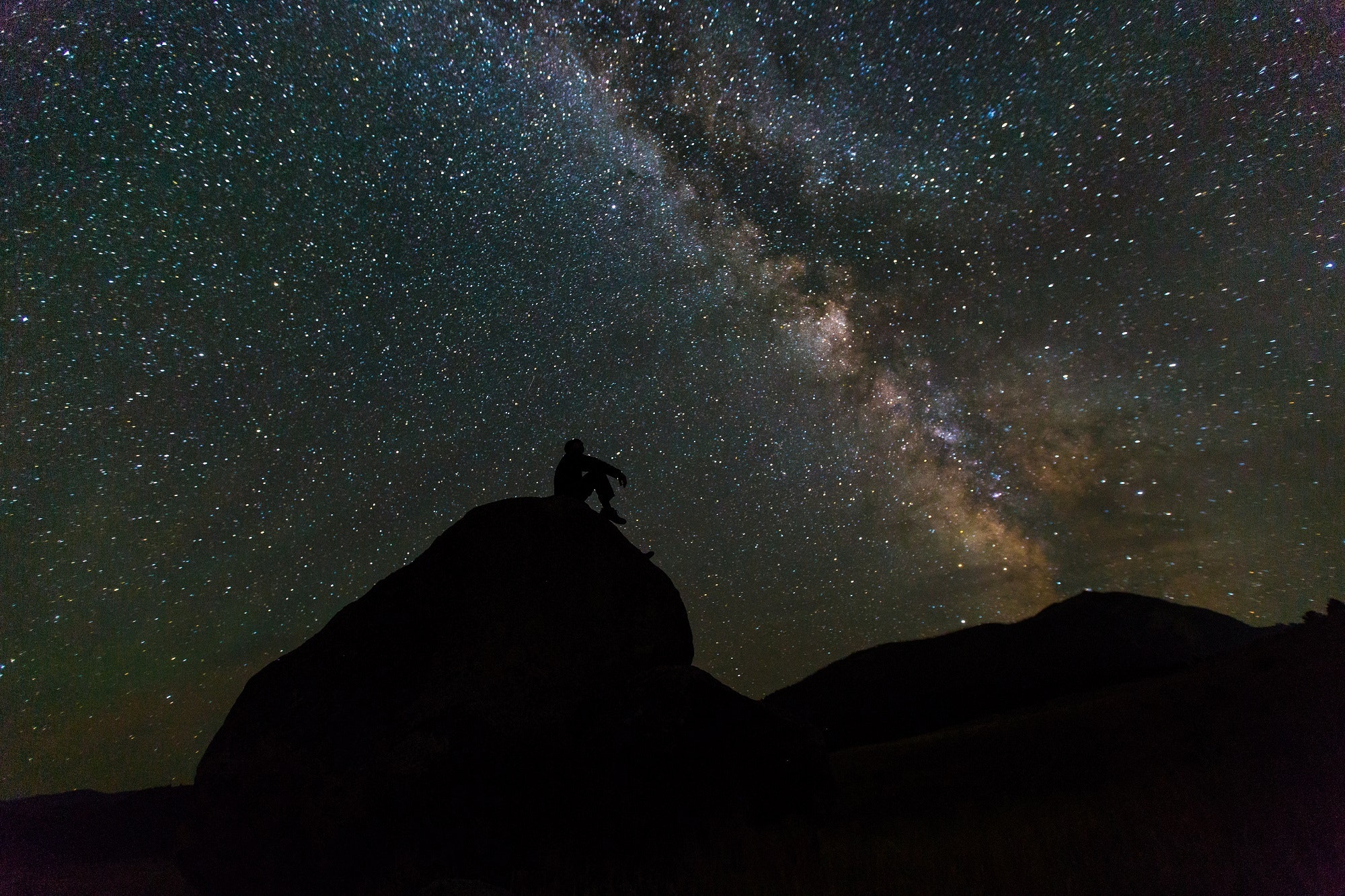 A silhouette of a person sits on a rock, gazing up at an incredible number of stars in an inky black sky.