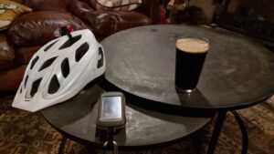 A round coffee table in front of a red leather sofa contains a cycle helmet, a GPS receiver (the first two digits of the time - 19 - are visible on its screen), and a pint of dark-coloured beer.