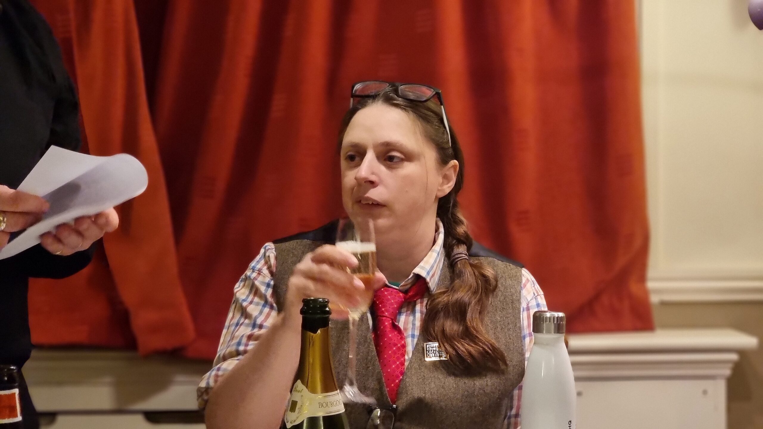 A woman dressed in a tweed waistcoat, glasses perched atop her head, sips a glass of champagne.