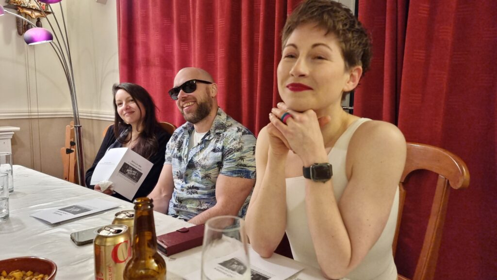 Three people sit alongside a dining table: a woman with dark hair, holding her play notes, a man wearing sunglasses and a Hawaiian shirt, grinning, and a short-haired woman in a conforming white off-the-shoulder top and bright red lipstick. There are beer bottles, champagne flutes, and Coke cans on the table.