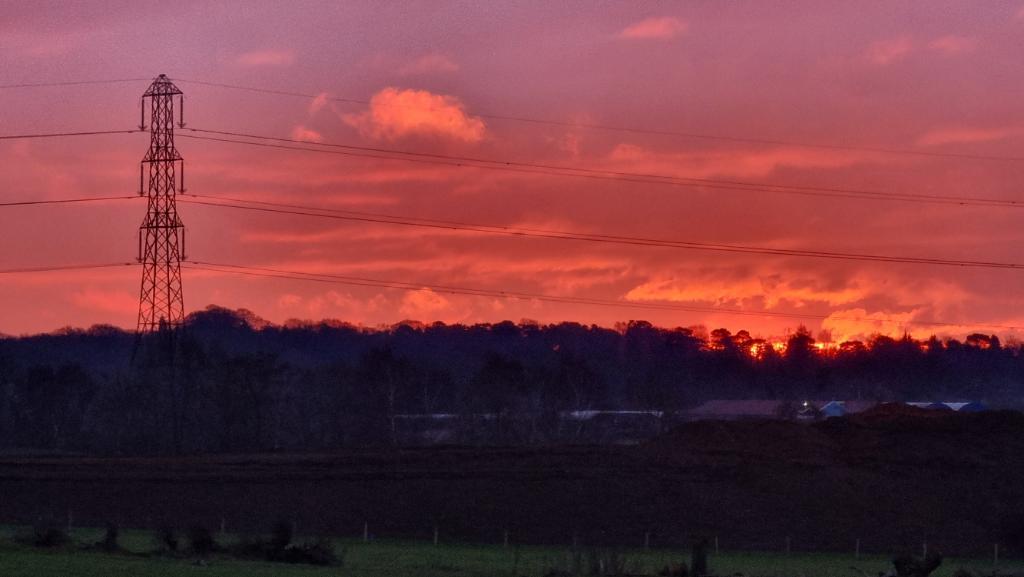 A deep red sunrise behind some power lines, over a green field and a shadowy hill.