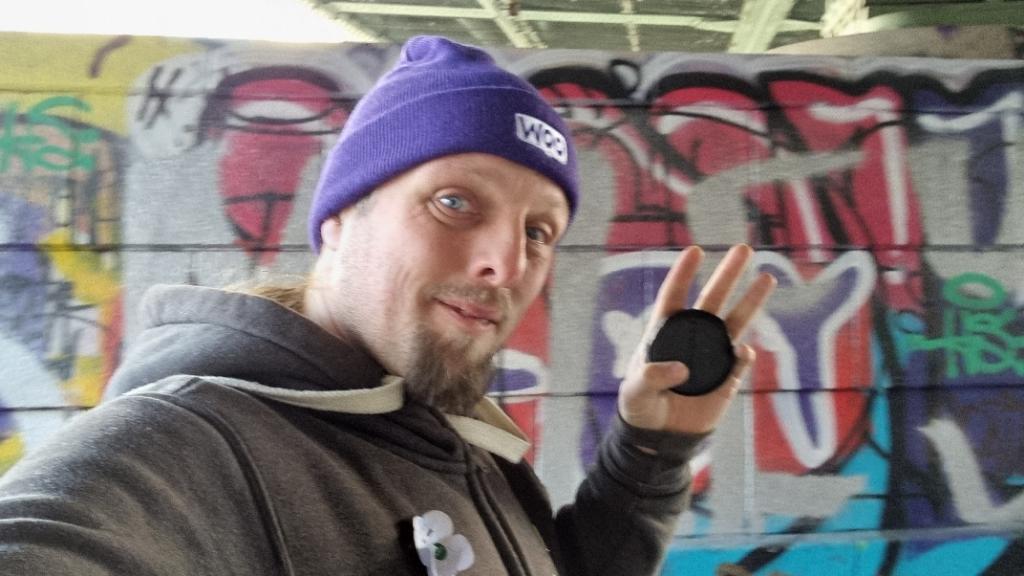 Dan, wearing a grey hoodie and a purple "Woo" hat, holds a black puck-shaped geocache. Behind him, a concrete bridge support pillar is decorated with colourful graffifi.