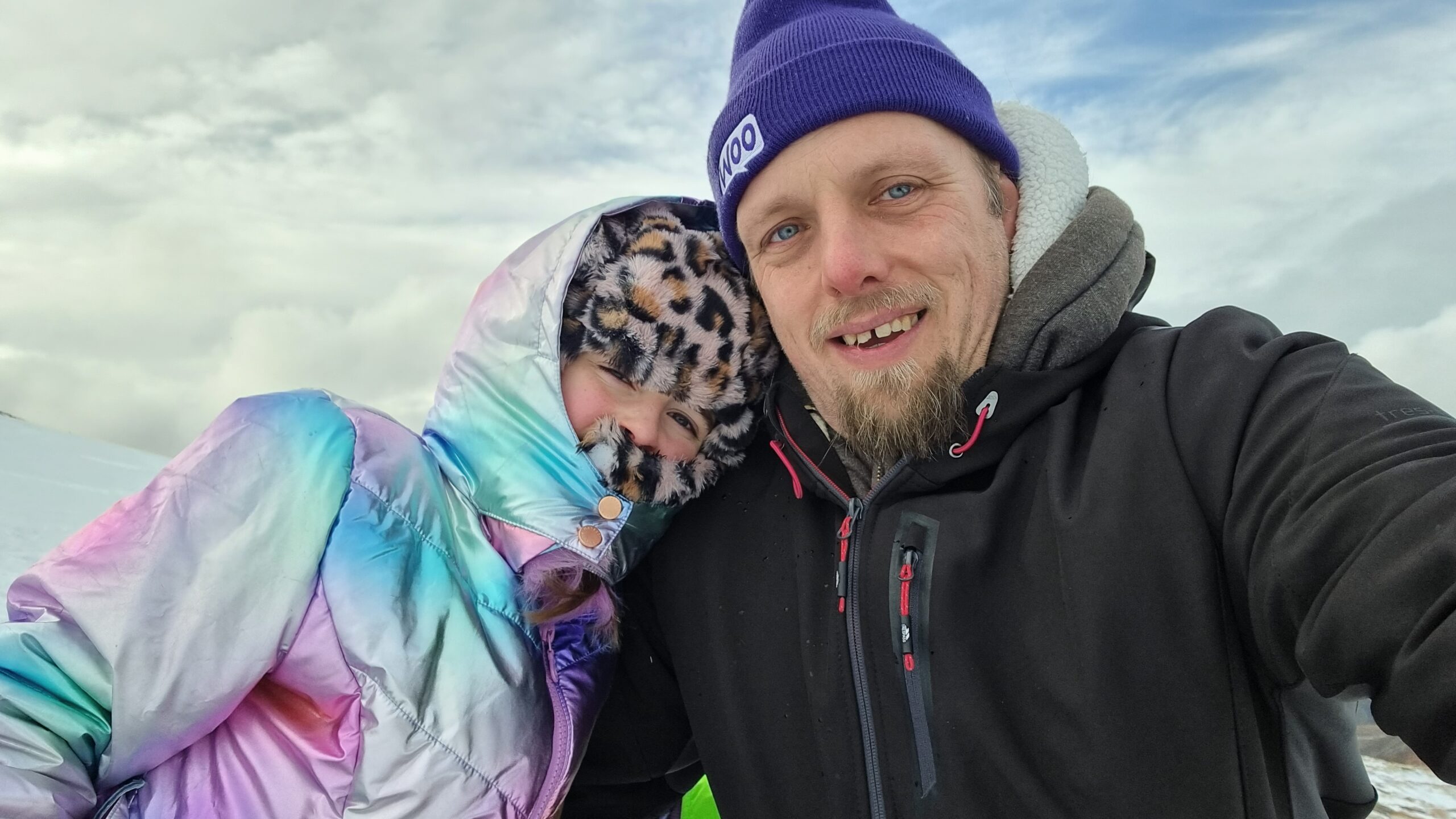 Dan, wearing a warm weatherproof black jacket and a purple "Woo" woolen hat, alongside a 9-year-old girl wrapped up in a faux-leapordskin hat and an iridescent coat, against a snowy hillside with rolling clouds.