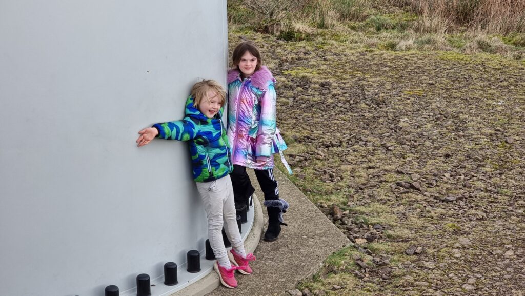A smiling boy and girl in brightly-coloured coats hug the bottom of a very large wind turbine stem.