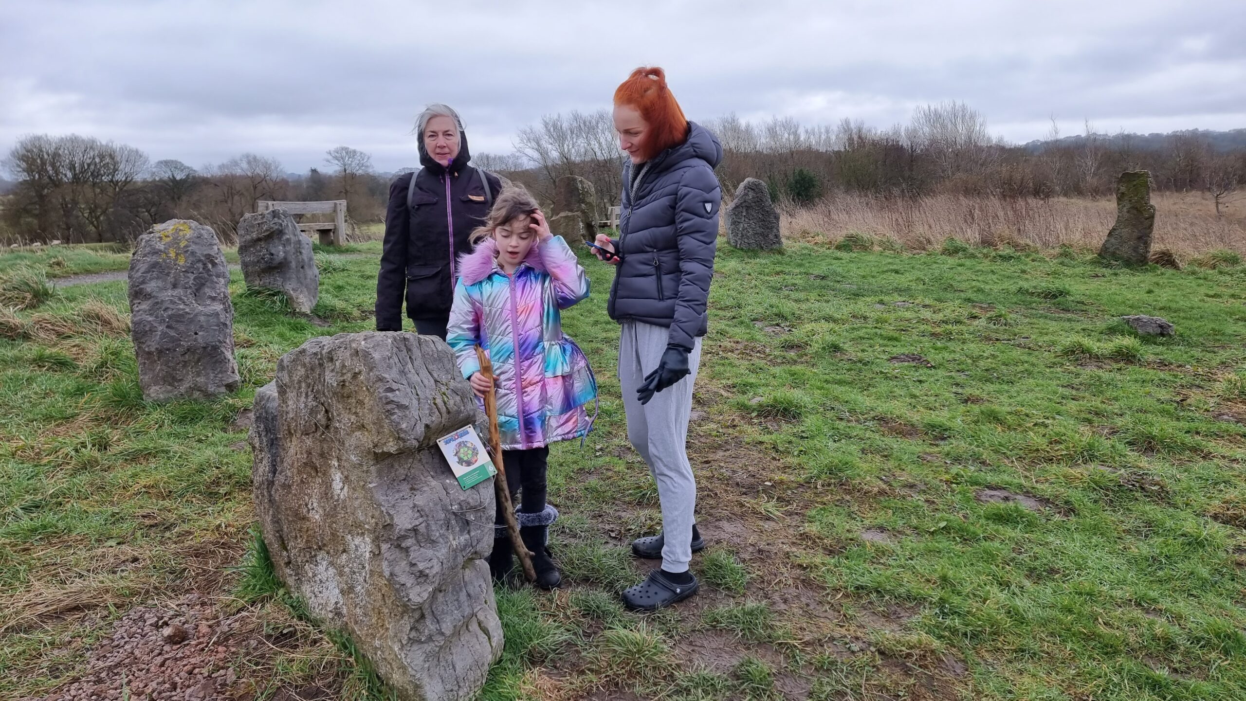 In a grassy field, a 9-year-old girl in a bright coat, accompanied by two women, examines a standing rock at the edge of a small stone circle.