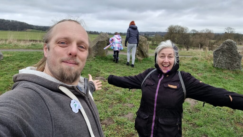 Dan and his mother stand in a stone circle, their arms spread wide and smiles on their faces.