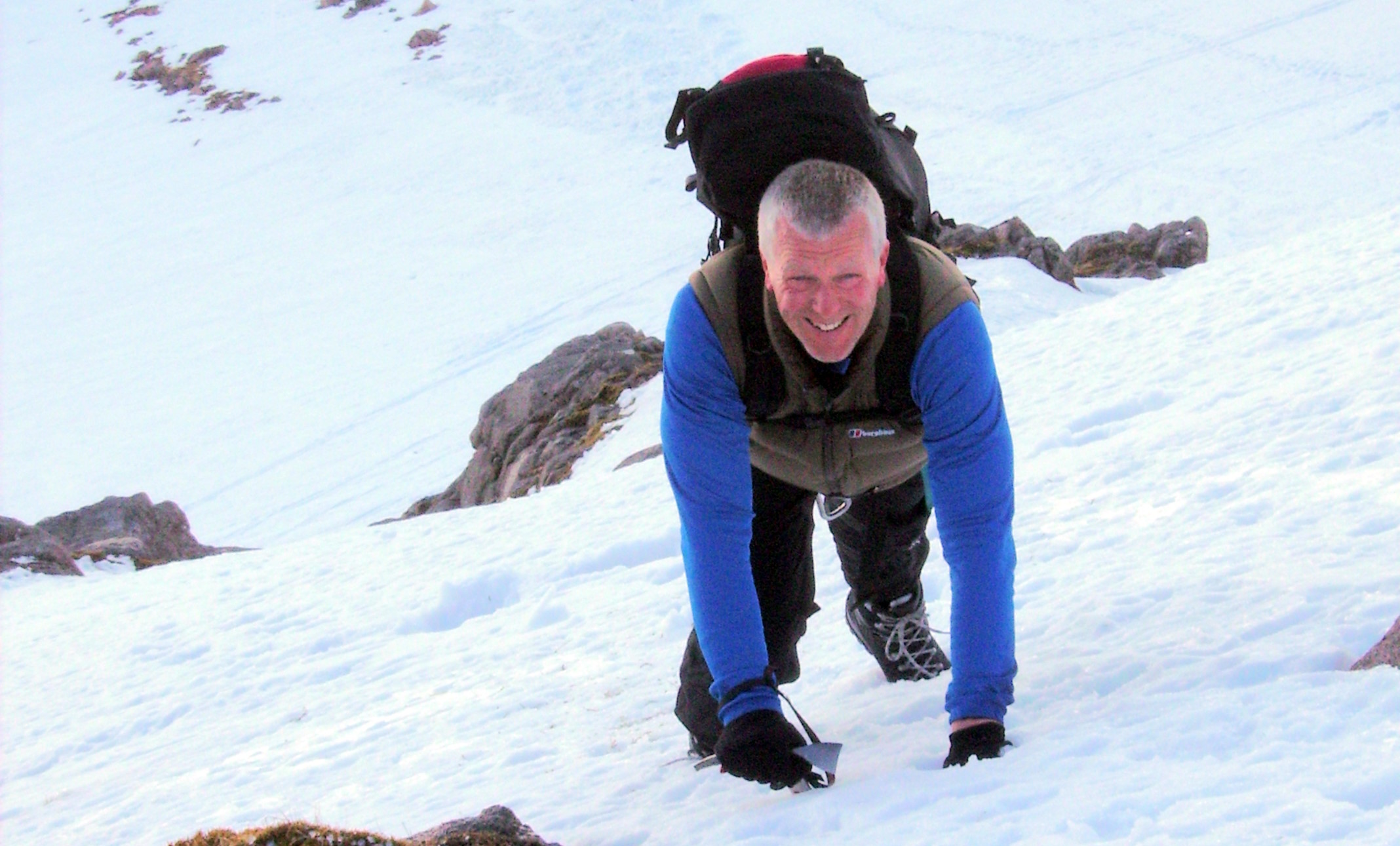 Seen from above, a man in his 50s wearing a large backpack uses mini ice axes to scramble up a steep hillside of powdered snow and rocks.